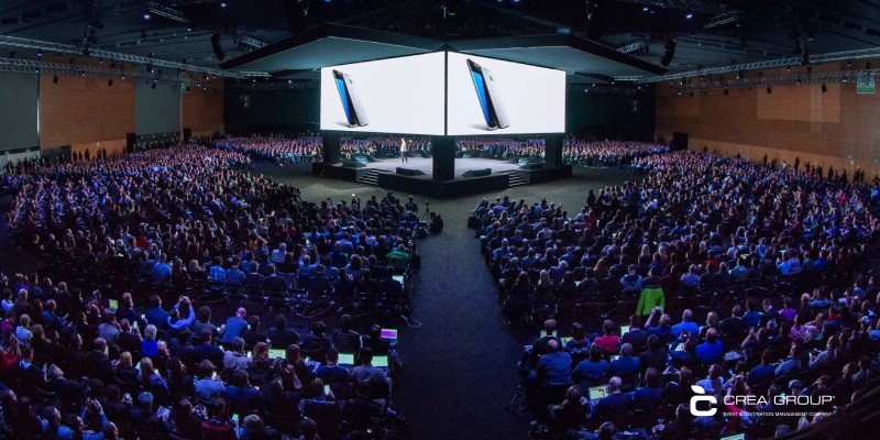 Gran audiencia en una sala de conferencias observando a un ponente en el escenario con pantallas gigantes mostrando un producto smartphone